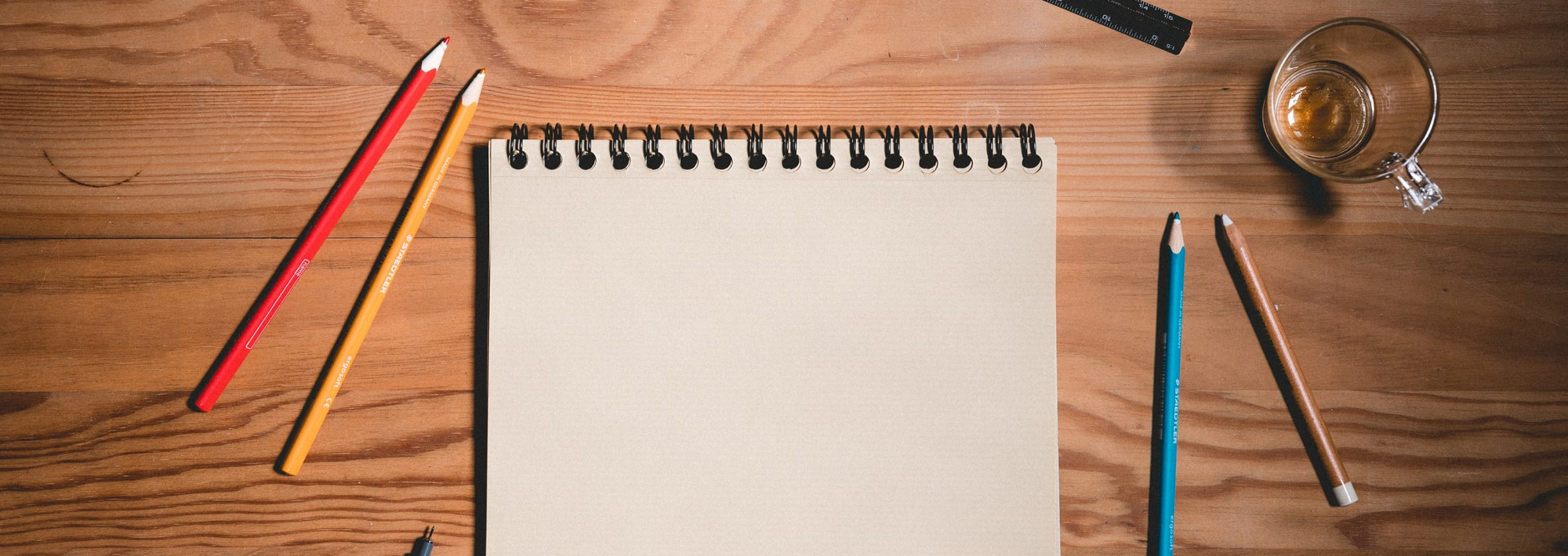 A notebook on a wooden table, with pencils around the notebook. The page is blank.