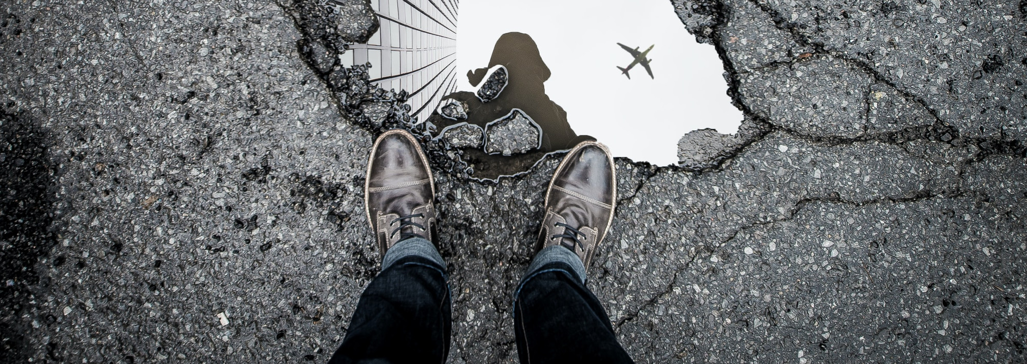 A person standing on a street, looking down at a puddle on the ground. The sky is reflected in the puddle.