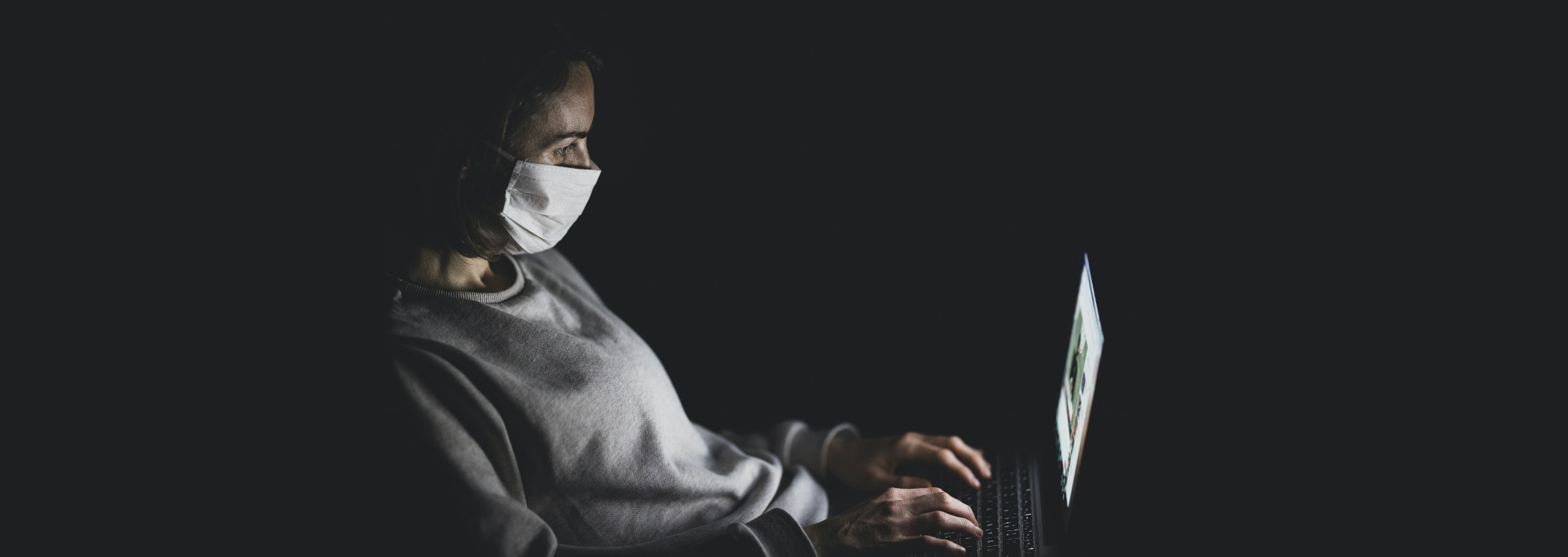 A woman wearing a face mask in a dark room, lit only by a laptop screen