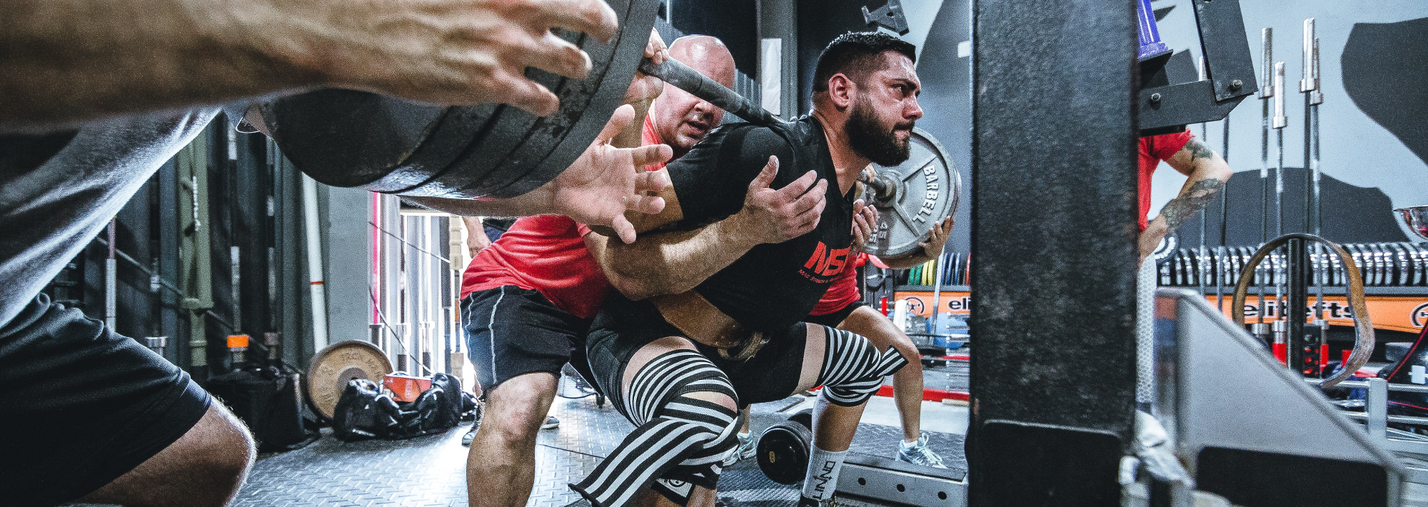 A man squatting with a barbell. Multiple people assist him in case the weight is too much.